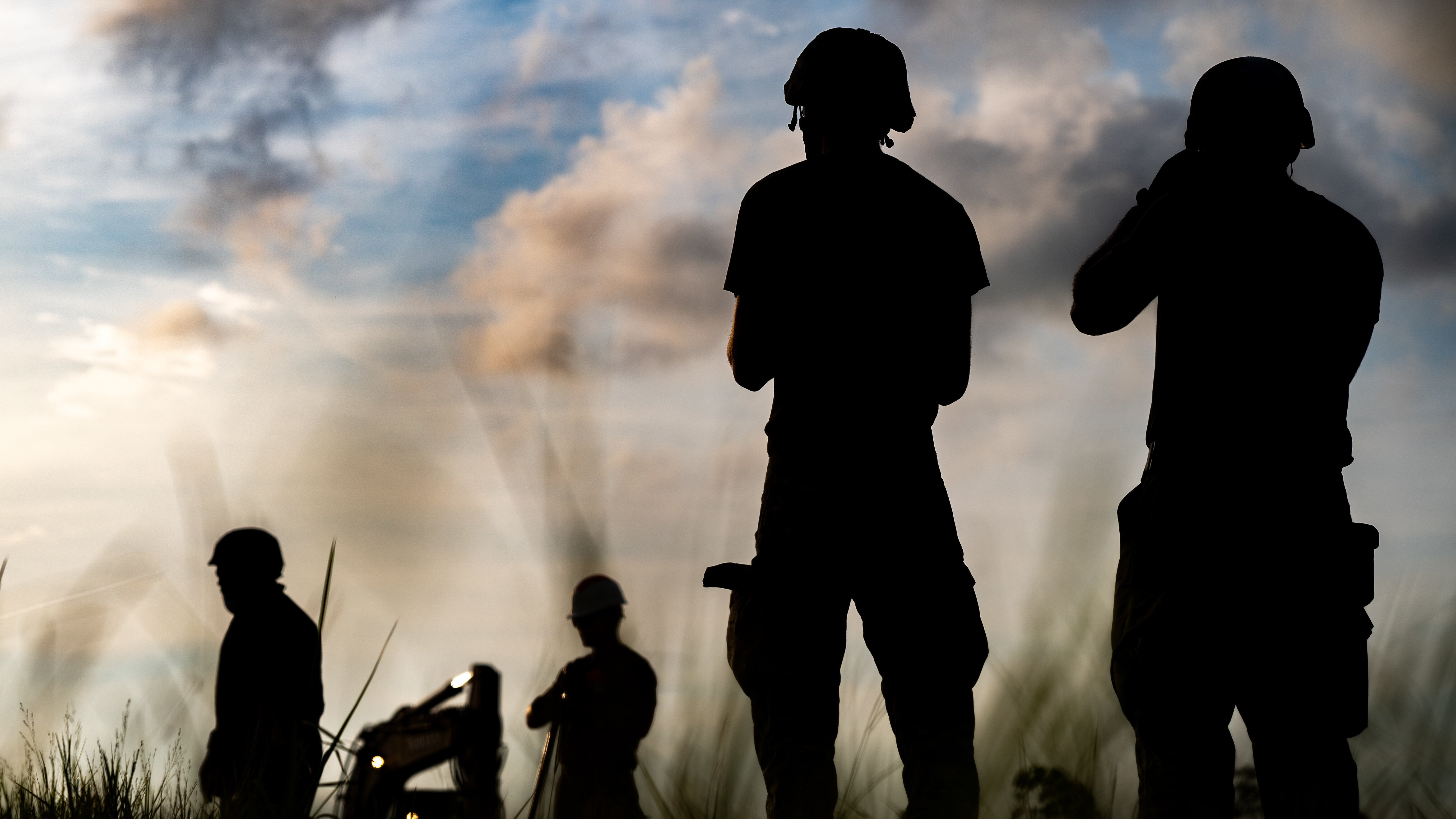silhouette of four airmen standing in field with equipment, smoke and clouds in blue sky