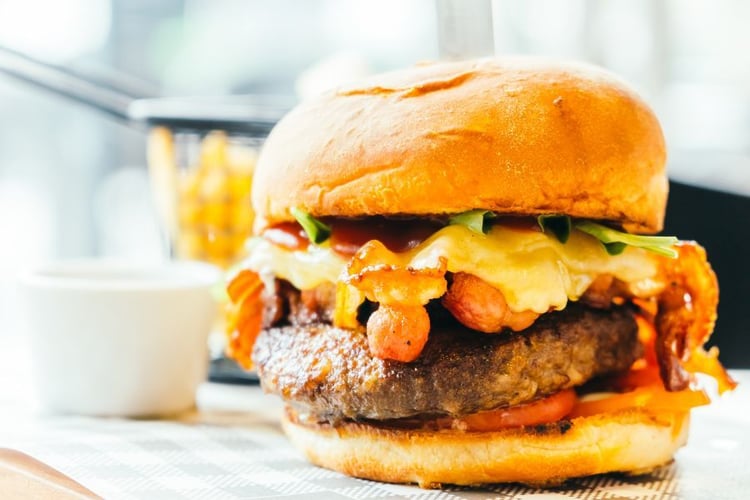 close up of hamburger with cheese, bacon, lettuce, tomato against restaurant backgrop and white cup