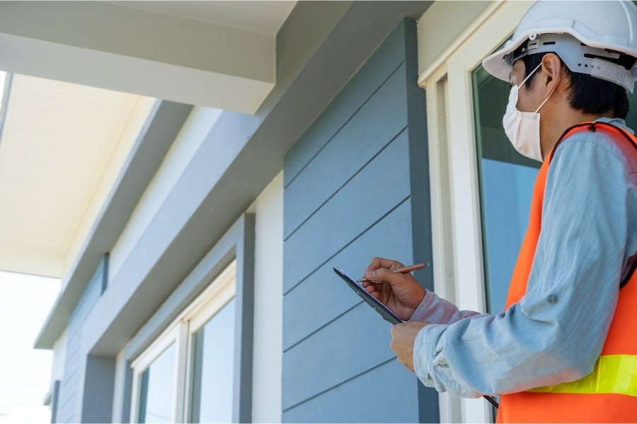 man in mask and orange vest inspecting blue home and writing notes on a clipboard