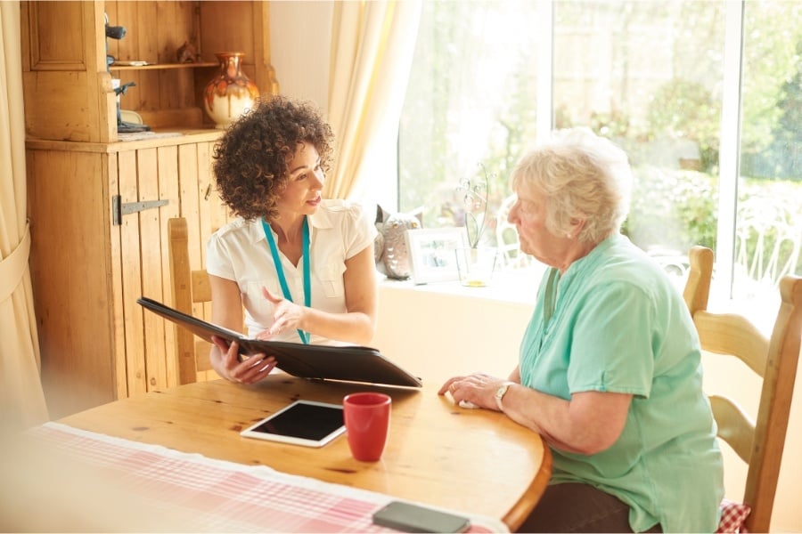 a young woman going over a tenant agreement with an elderly woman while they both sit on chairs at a kitchen table with window behind them