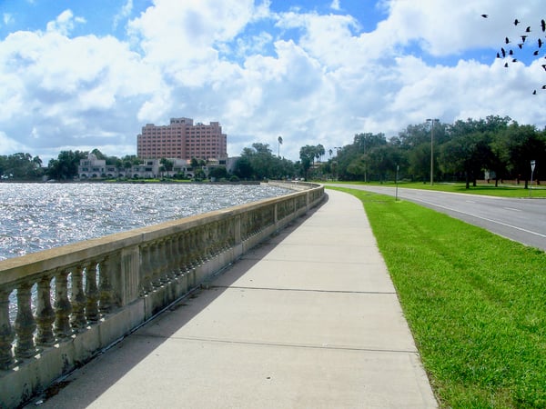 This is an image of Bayshore Blvd in Tampa, FL, near MacDill AFB. There is a cement path along the water with grass on the right. There are trees and a multiple story hotel in the background. The sky is blue with fluffy clouds.