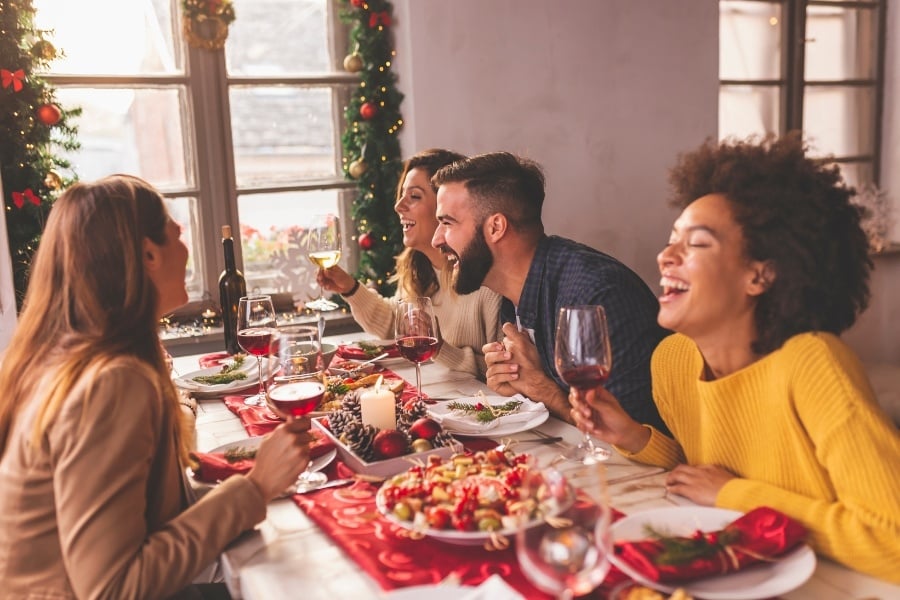 man with beard and women gathered around table with holiday food, candle, and wine with christmas garland and ornaments hanging behind. 