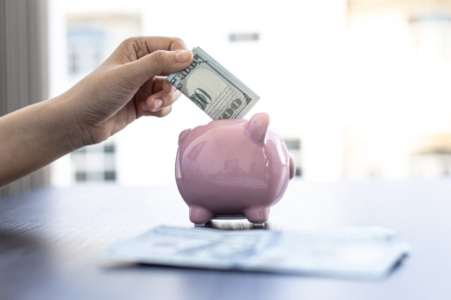 a person's hand putting one hundred dollar bill into pink piggy bank