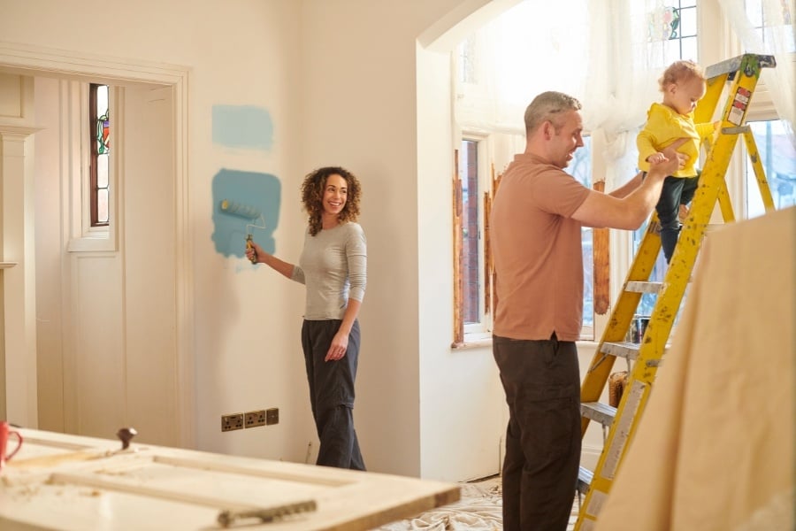 Smiling man and woman paint and balance toddler on yellow ladder while renovating house.