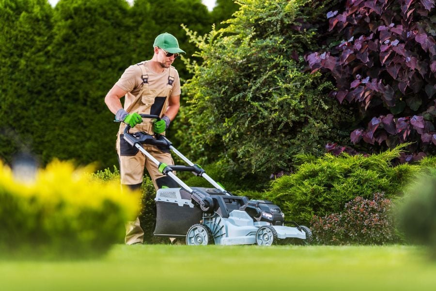Professional lawn service provider in tan utilities with green hat and gloves pushes mower against green lawn with lush trees around.