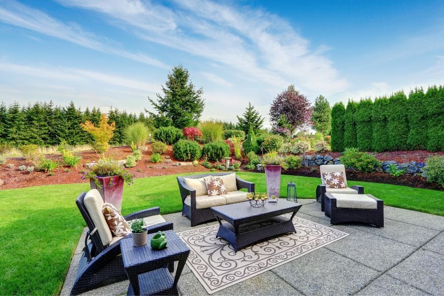 Extended patio with area rug, loveseat, coffee table and two chairs in green lawn with beautiful landscaping lining the yard.
