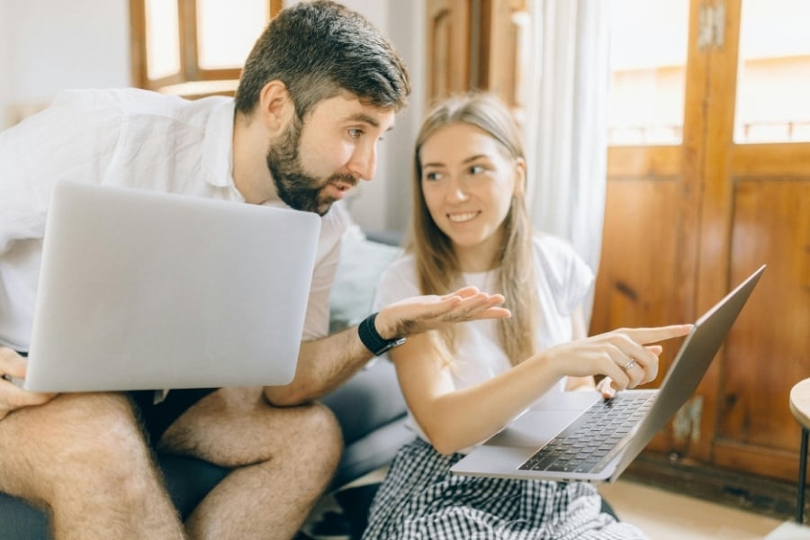Couple in white shirts sit on couch and floor and compare home listings on each other's laptops. 