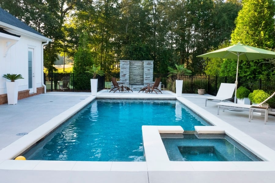 In-ground swimming pool in backyard with white loungers, umbrella, and line of trees around. 