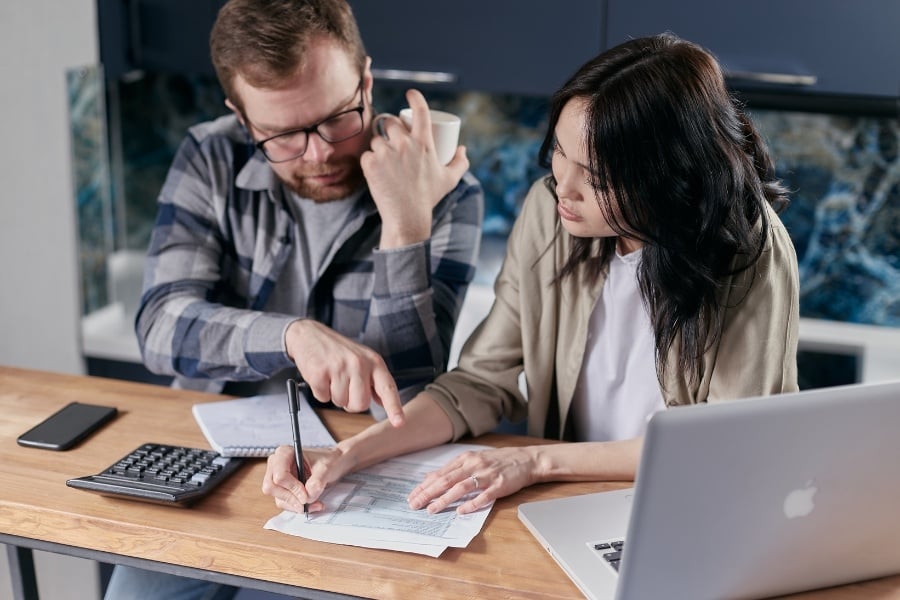 Man with beard and glasses sitting with woman with long dark hair pointing at papers next to computer and calculator 