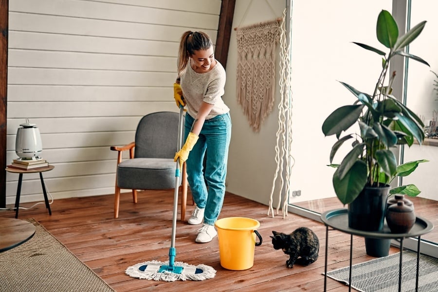 Woman with ponytail, jeans, and white sneakers mopping wood floor with yellow gloves and bucket. Cat sits next to bucket and room is neutral with large green plant 