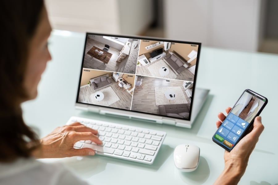 Woman view smart home security cameras from app on phone and views from around the house on the computer in front of her. 