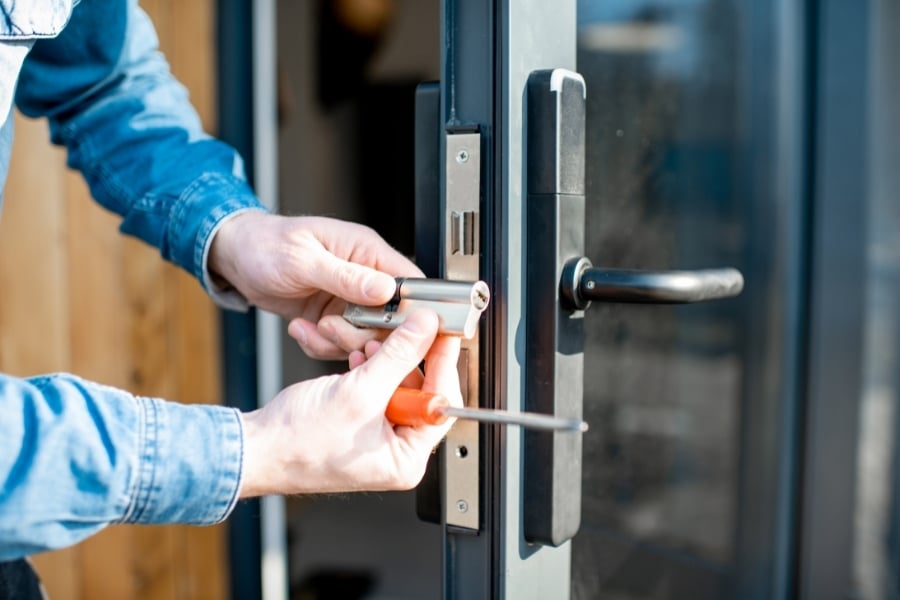 Man in denim long-sleeve shirt changes out lock on front door with orange screwdriver. 