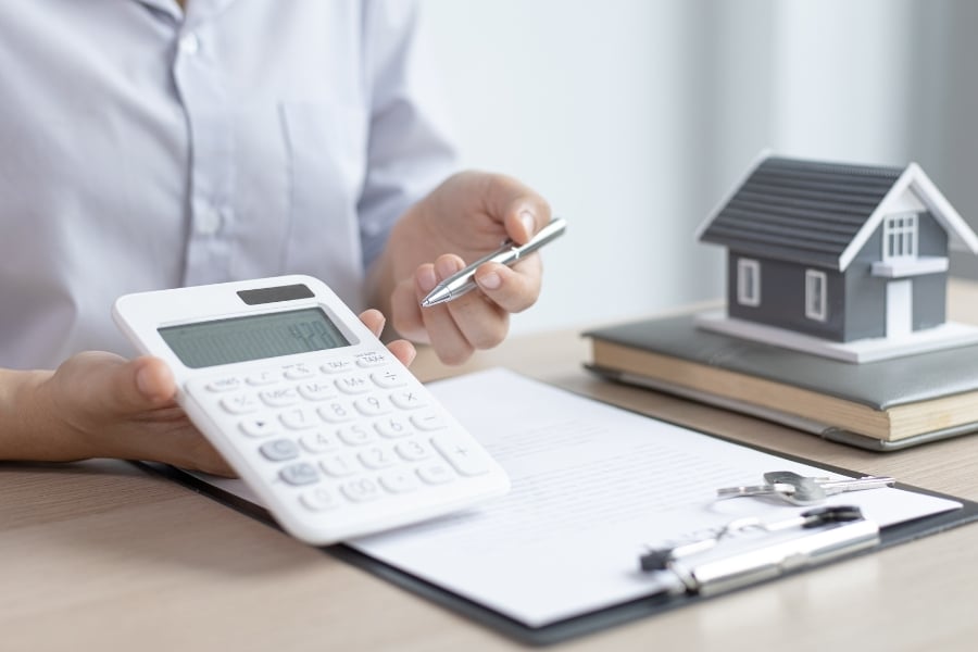 Man in button down with gray model house and notebook holds calculator above clipboard and points to number display with pen. 