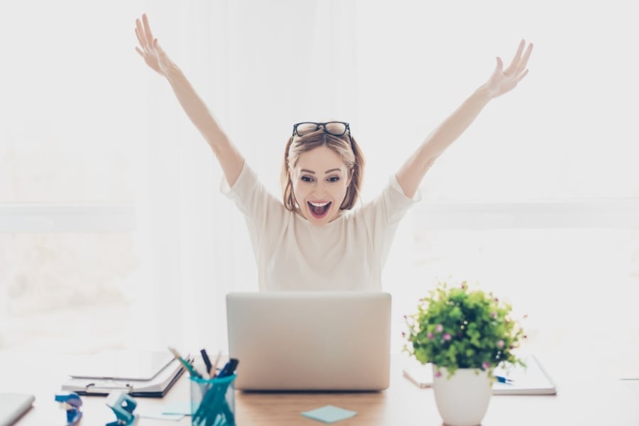 Woman with blonde hair, glasses on her head, and white shirt raises arm in excitement looking a computer. cup of pens and a plant sit on her desk