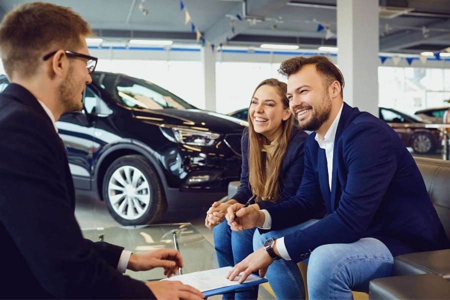 Man with beard and girl with long hair sit down with man wearing glasses to sign paperwork and buy car, black car sit in the background