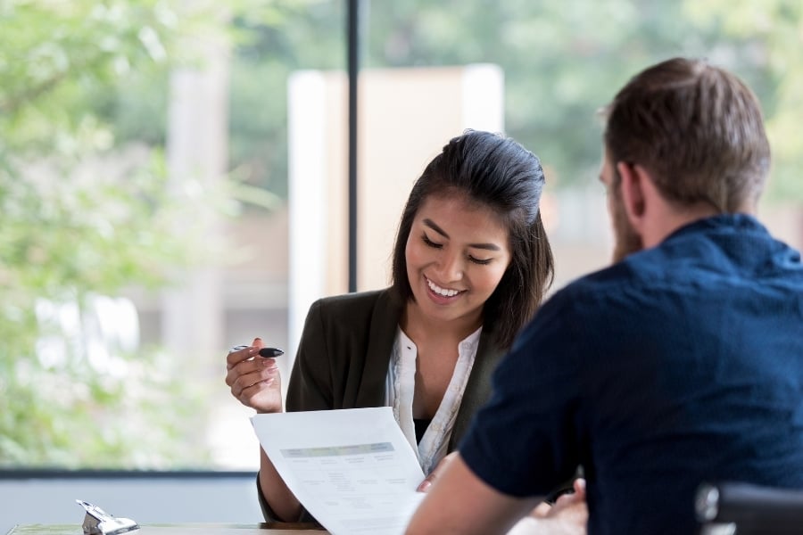 Woman in blazer sitting in front of large windows smiling and pointing at paperwork in hand to man in blue shirt with back to camera. 