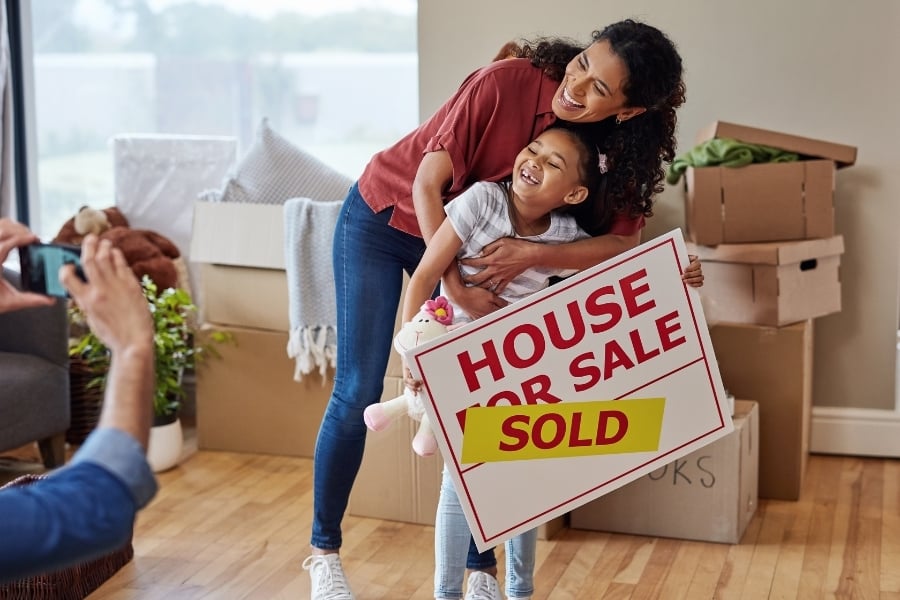 Dad taking photo of mom with arms wrapped around daughter who's holding a House for Sale sign marked sold while smiling in house with boxes around. 
