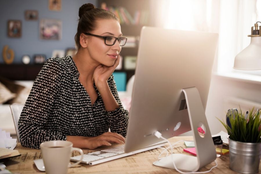 Woman wearing glasses rests head on hand while looking at computer on desk next to cup of coffee.