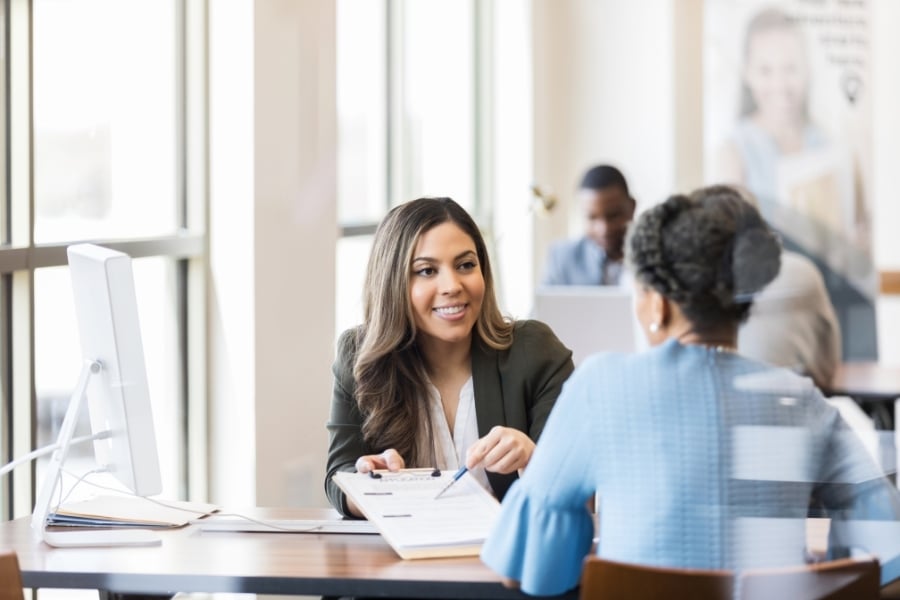 Woman in blue sits across from female loan officer wearing a blazer and white shirt smiling at client while pointing to paperwork on clipboard. 