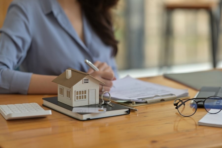Female loan officer in blue button down writes on paper sitting on clipboard with small model house, keys, and glasses next to her on the wooden desk. 