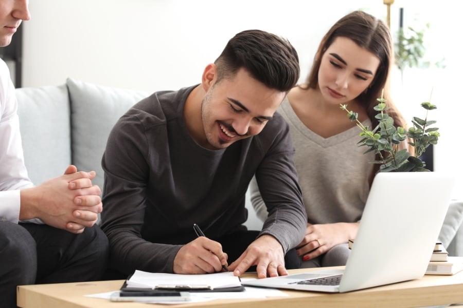 Man in dark shirt smiles as he signs paperwork by computer on coffee table with real estate professional on one side and woman on the other side watching him. 