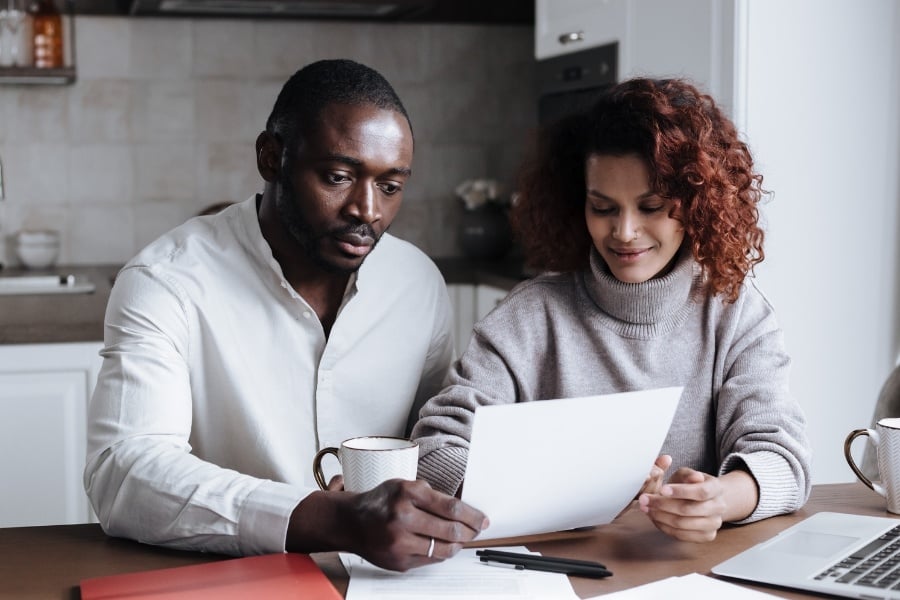 Photo of couple sitting at table looking at papers in front of computer. 