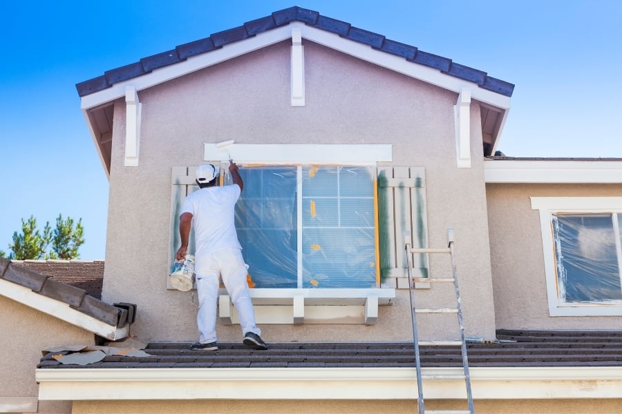 House painter in all white stands overhang from first floor to paint trim around second-story window with ladder nearby. 