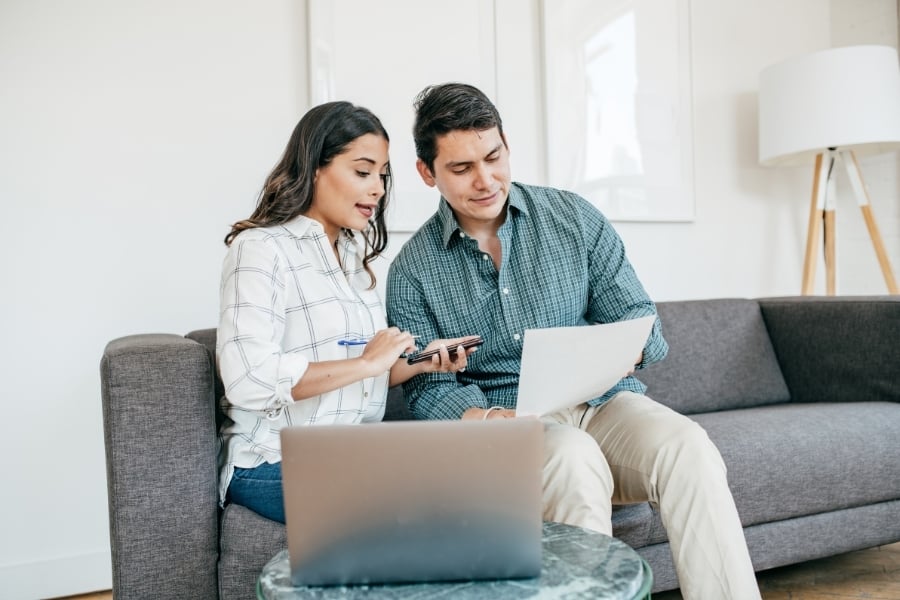 Man and woman wearing button-down shirt sit on gray couch reviewing papers and phone in front of computer.