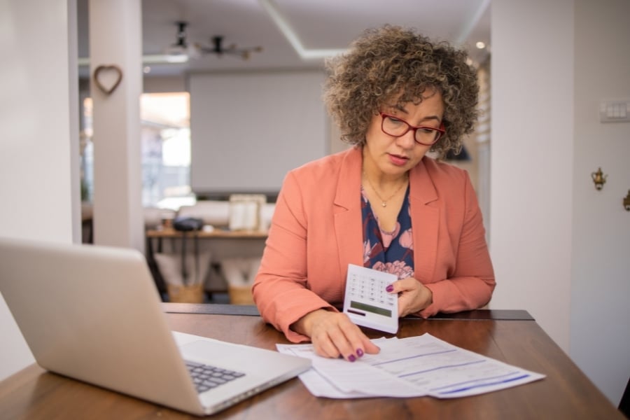 Woman wearing orange blazer and red glasses sits with calculator in hand and looks at papers sitting in front of her next to computer. 
