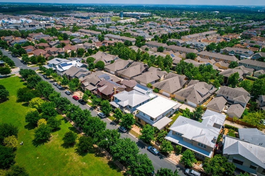 Aerial view of suburban neighborhood with tree-lined roads, cars parked, and a grassy area nearby. 