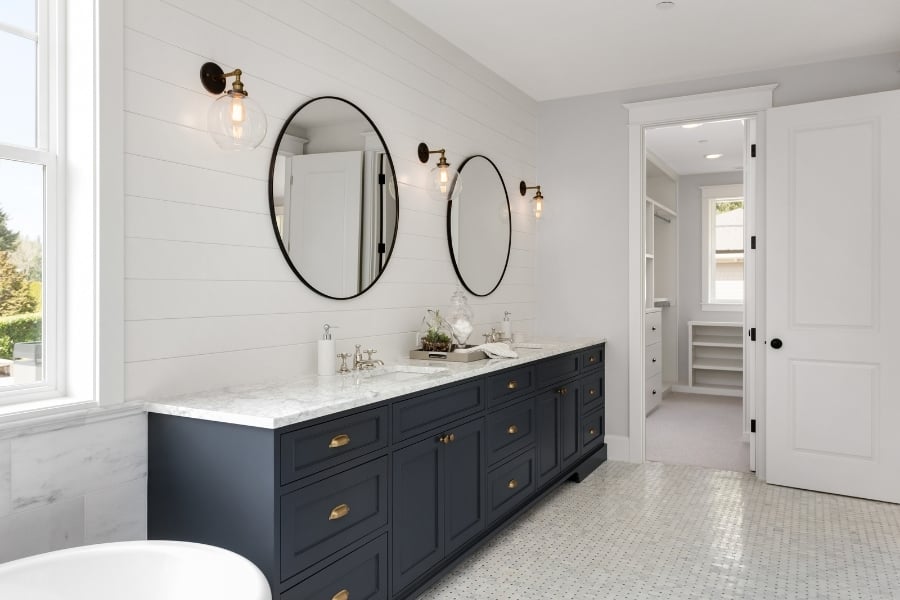 Large bathroom with navy blue double sink vanity, black oval mirrors, a garden tub, white tile flooring, and an open door leading to walk-in closet with a window.