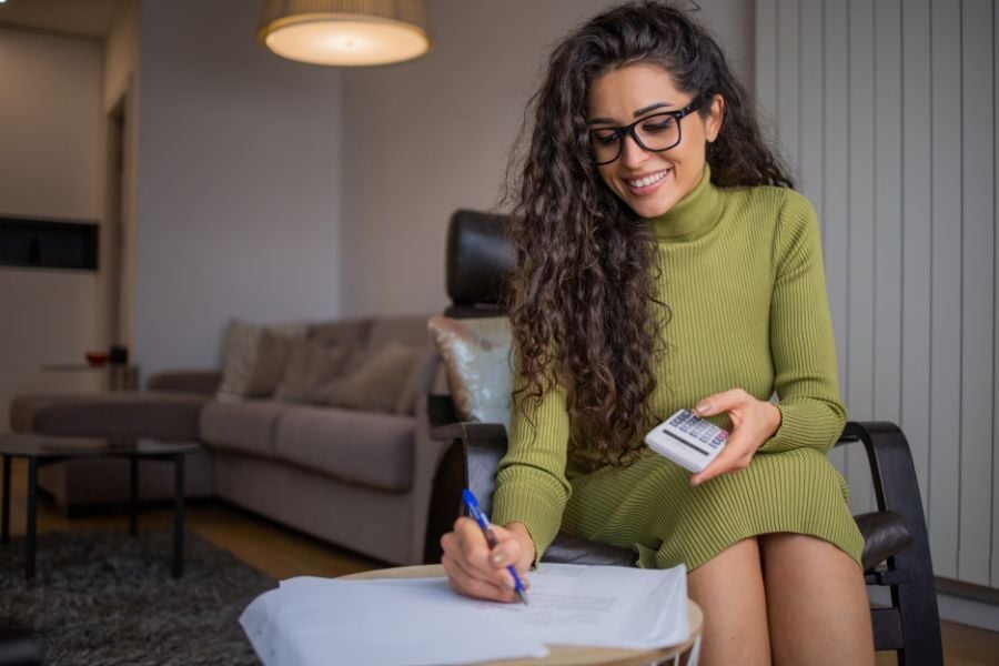 Woman in green sweater dress holding calculator in hand and writes on papers sitting on small side table 