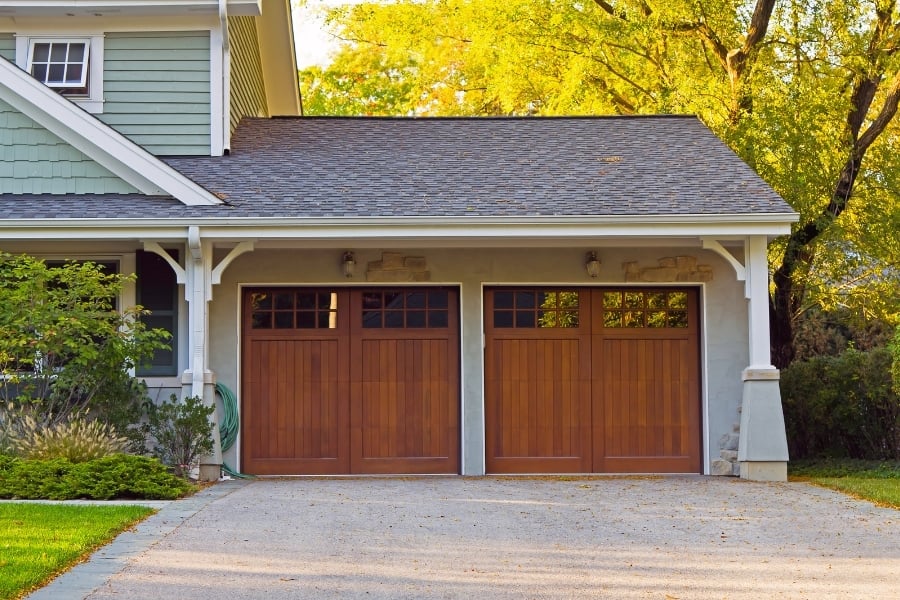House with sage green siding and white trim has beautiful wooden garage doors with windows that are centered in the frame with lush, mature trees positioned behind. 