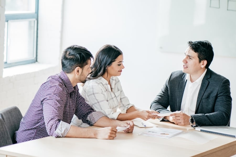 Man in purple button down sits with woman in plaid button down across from man in dark suit negotiating over paperwork in front of them on the table. 