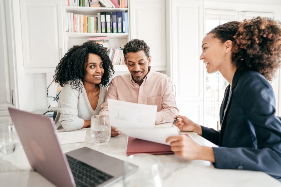 Man and woman look over papers as they meet with female professional sitting at white counter with laptop. 