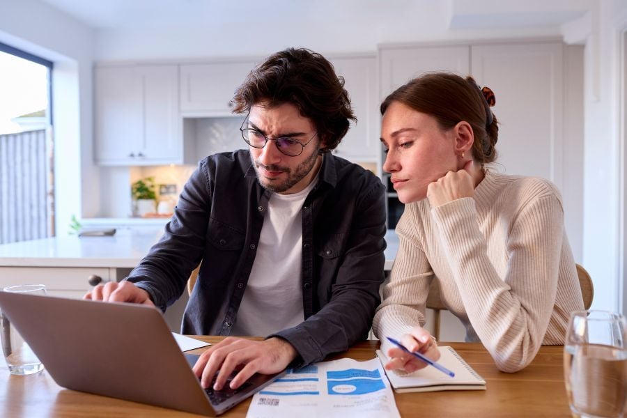 Man in glasses points at computer to show the woman in tan who's sitting next to him with her head leaning on her hand. 