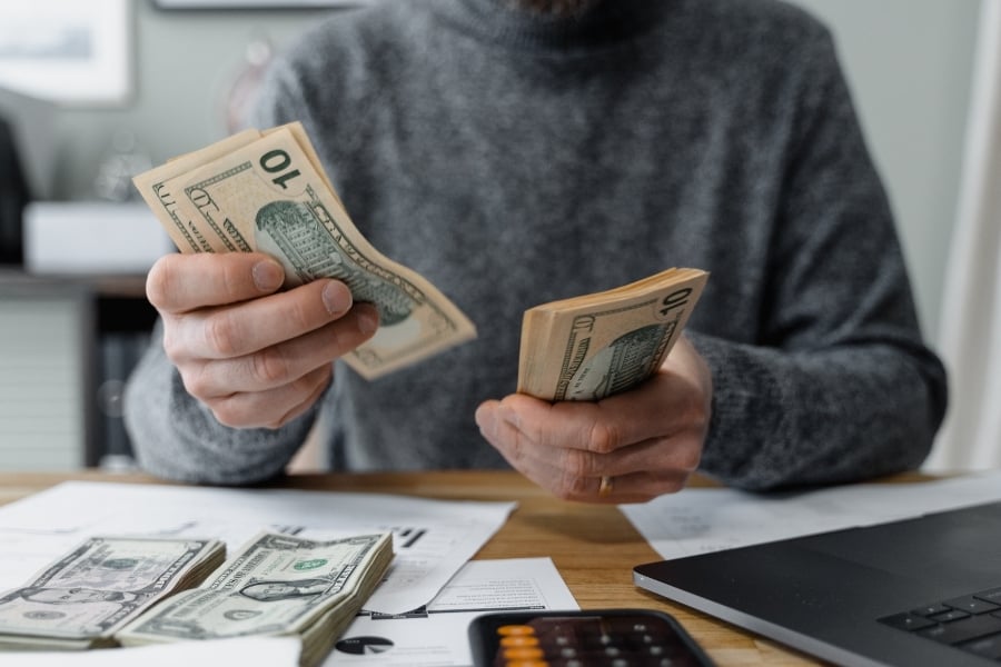 Man in gray sweater counts cash on wooden table with calculator and computer next to him. 