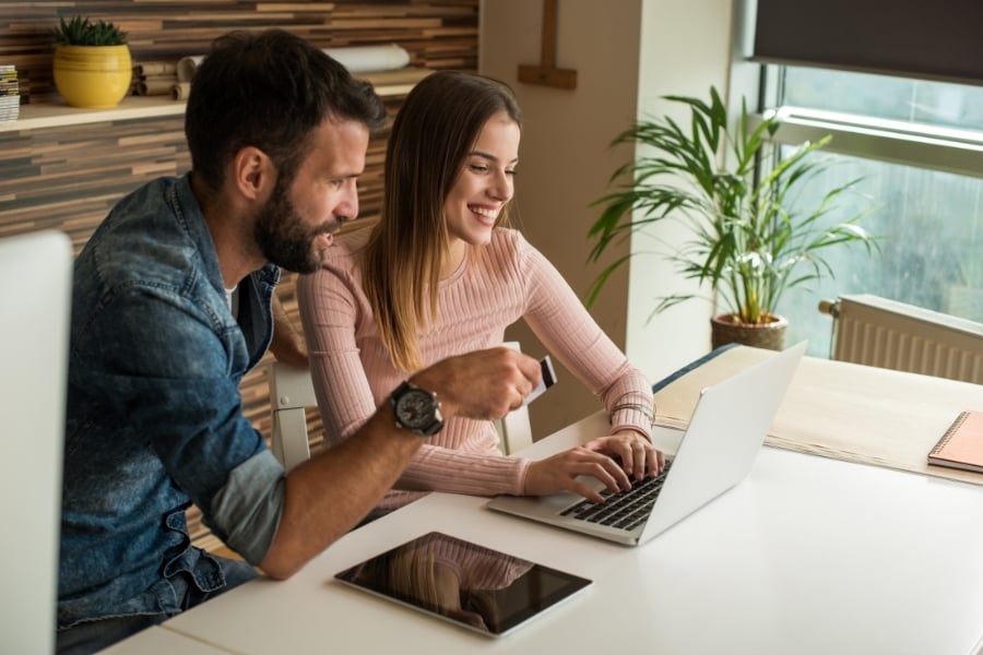Man in denim shirt sitting with woman in pink shirt smiling and pointing at computer on table in warm-toned room with plant sitting next to the window. 
