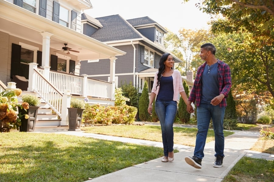 Man and woman in jeans and long sleeve shirts hold hands smiling while walking down the sidewalk in neighborhood with mature trees with the sun shining.
