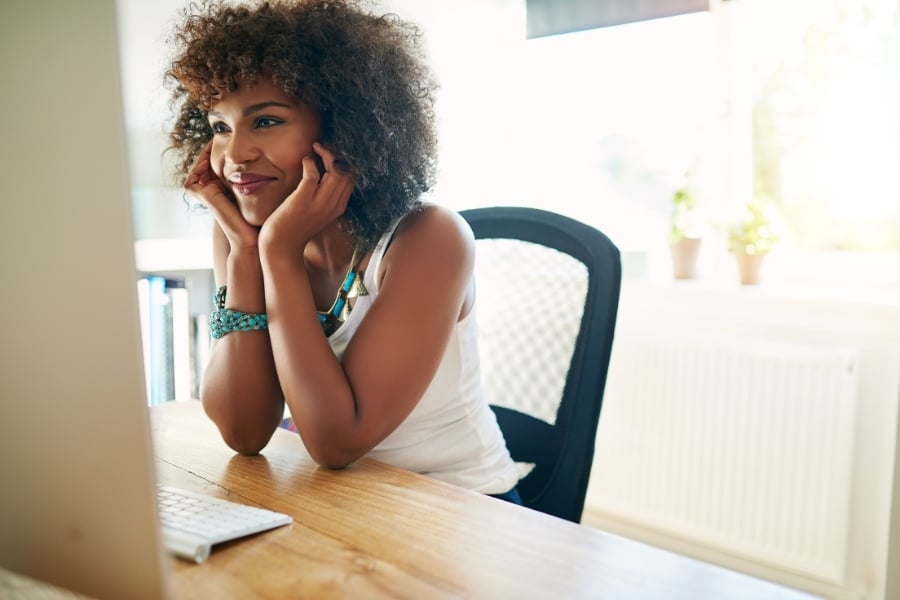 Woman in turquoise jewelry and white shirt smiling at computer with face in hands. 