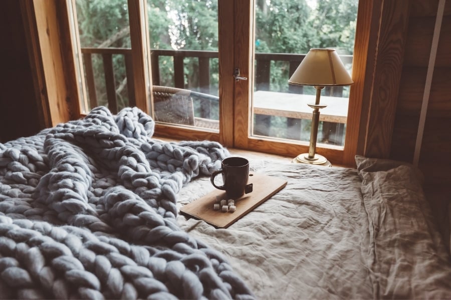 Cozy bedroom with wooden paneling and glass french doors leading to deck in woods. The bed has a thick, woven gray blanket laid across with cup of tea and sugar cubes sitting on tray. 