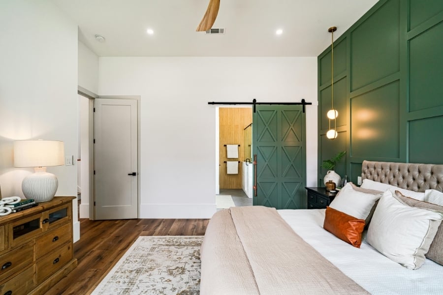 bed with pillows and duvet in modern bedroom with accent wall pained green, green painted door, throw pillows, rug, dresser with lamp on