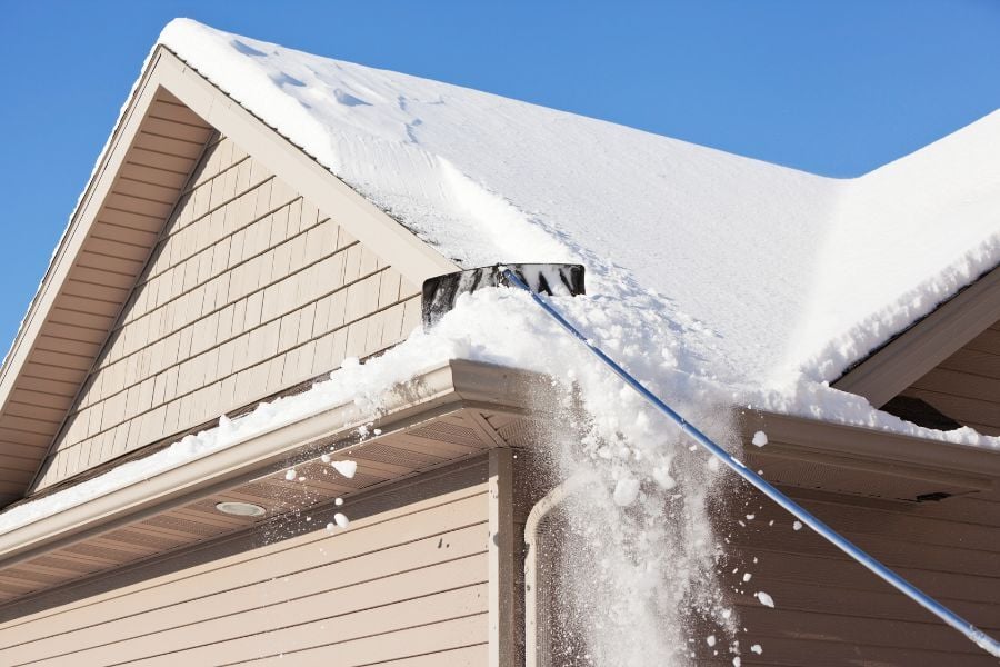 Long-handled shovel brushing snow off the roof of beige house with a blue sky above