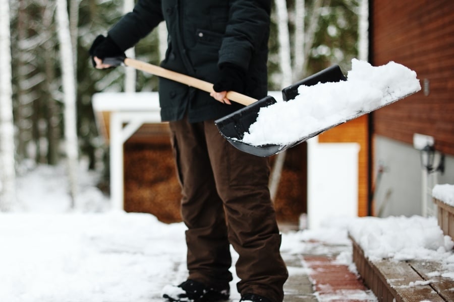 Person with black coat and brown pants holding shovel with snow outside wood paneled building and steps. 