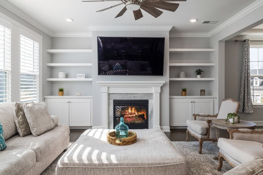 Beige living room with beige couch, chairs, ottoman, blue accents, large TV, bright window, built-in bookshelves, and fireplace burning 