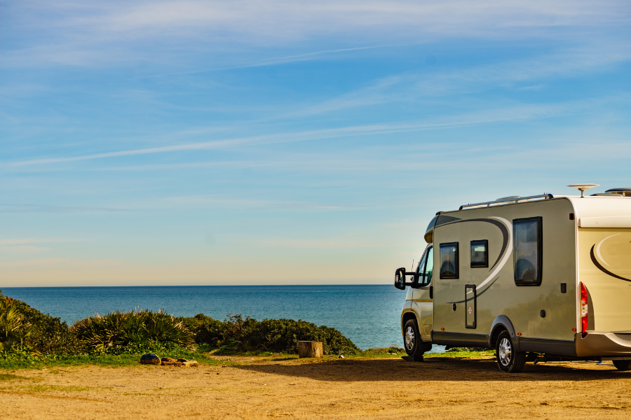 View of RV camper parked on sandy parking lot with bright blue ocean below. 