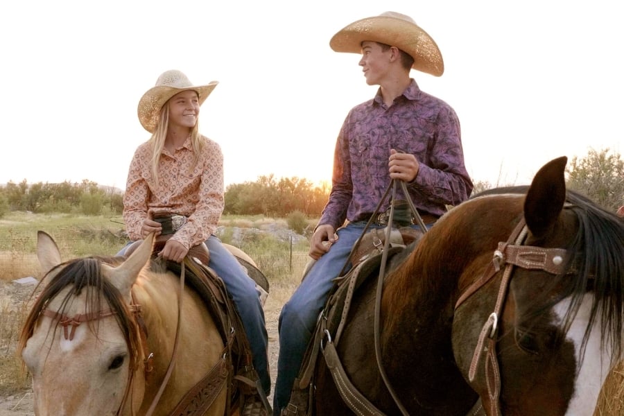 Boy and girl wearing western gear with cowboy hats, button-downs and denim jeans riding horseback smiling at each other. 