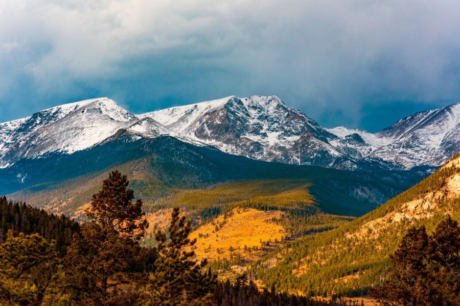 View of moody sky above snowcapped mountains with golden sunlight in Rocky Mountain National Park, Colorado