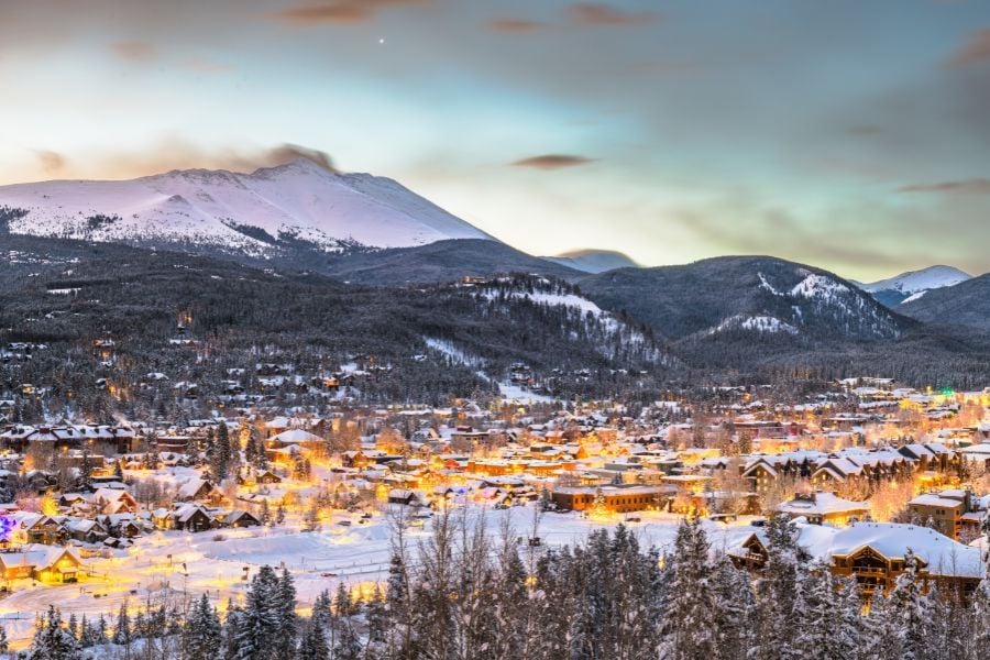 Aerial view of snowy Breckenridge, Colorado with faint sunlight over mountains and glowing lights of small city below.