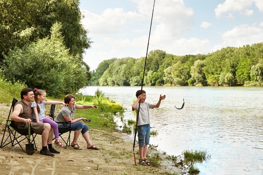 Family of four fishing along the river. Mom and dad sitting in chairs on river bank with daughter in lap while boy excitedly reaches for the fish at the end of his line. Everyone is smiling. 